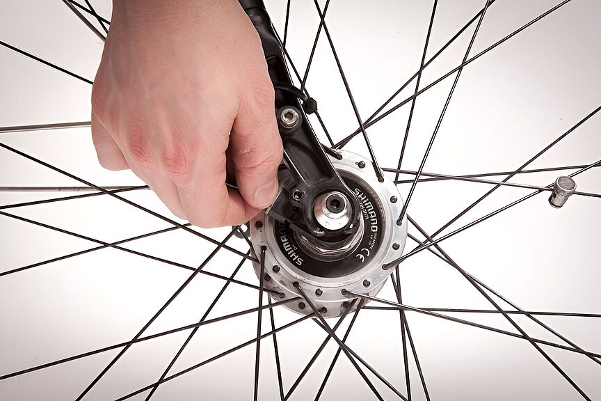 Feinjustierung an einer Fahrradnabe Nahaufnahme eines Fahrradlaufrads mit silbernen Speichen vor weißem Hintergrund. Eine Hand greift von oben in die Speichen und justiert mit einem schwarzen Werkzeug die Nabe in der Mitte des Rads. Rechts ist das Ventil des Reifens zu sehen.