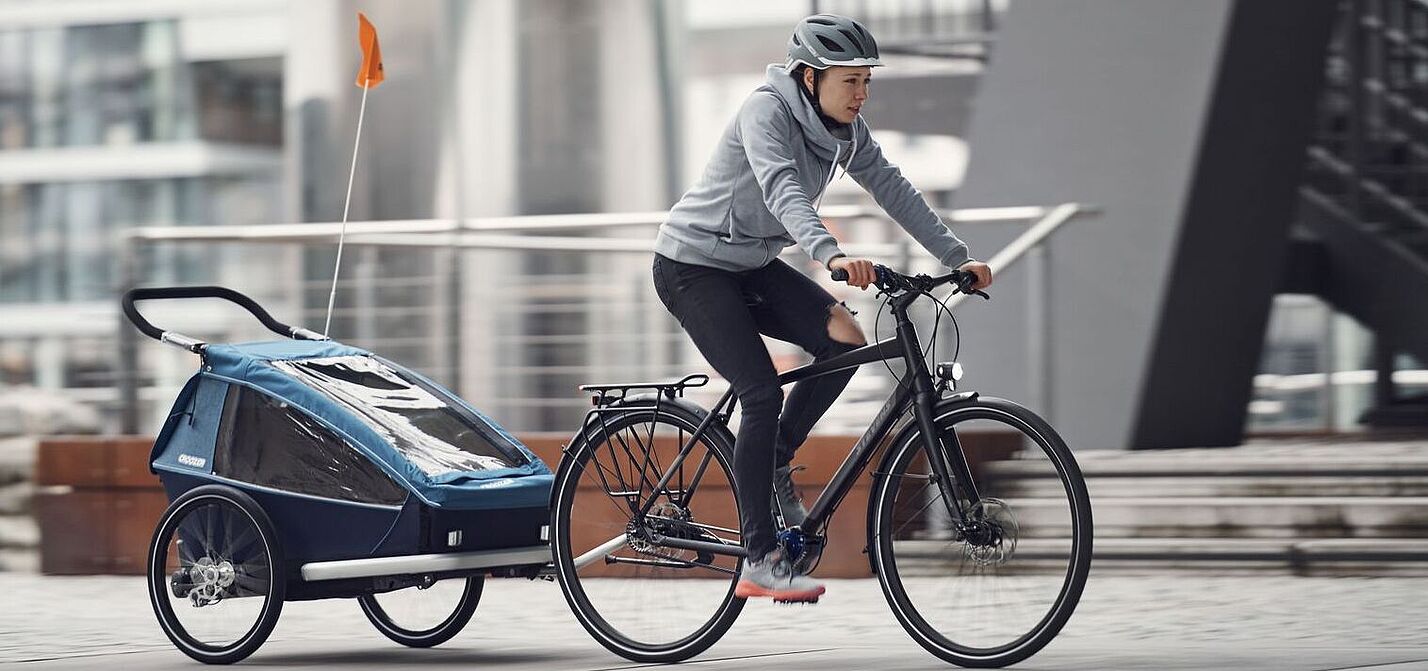 Das Fahren mit Kinderanhänger sollten Eltern üben. Eine Frau fährt auf einem Fahrrad mit einem Kinderanhänger.